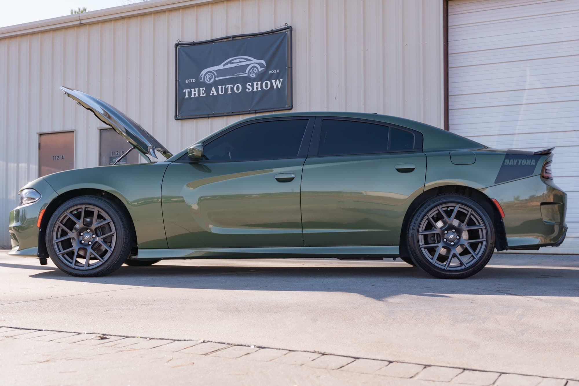 A green Dodge Charger with its hood raised, parked in front of The Auto Show building