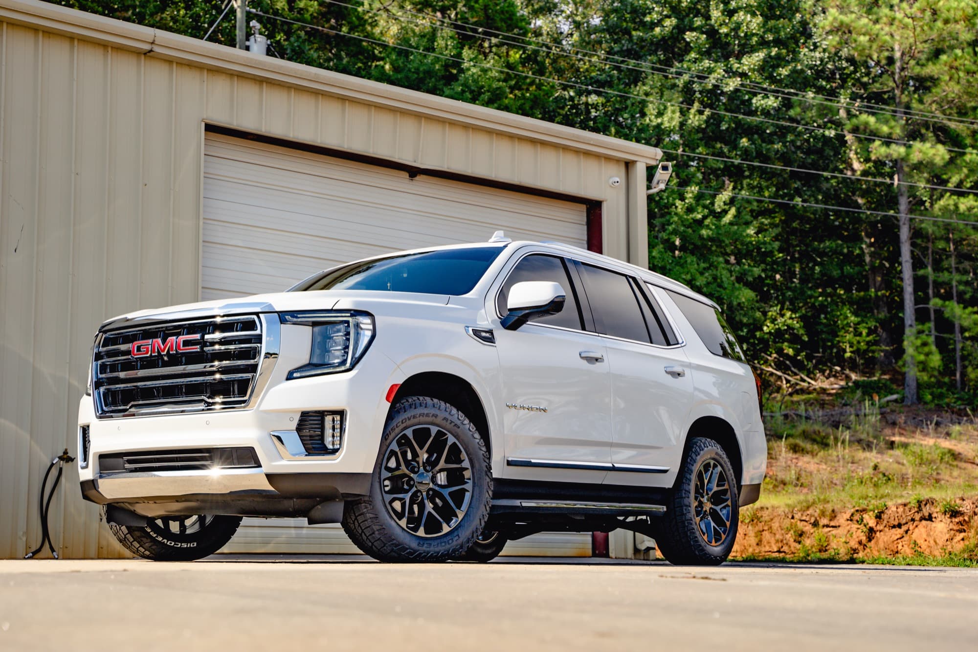 A white GMC Yukon with dark rear privacy glass parked outside a metal shop building