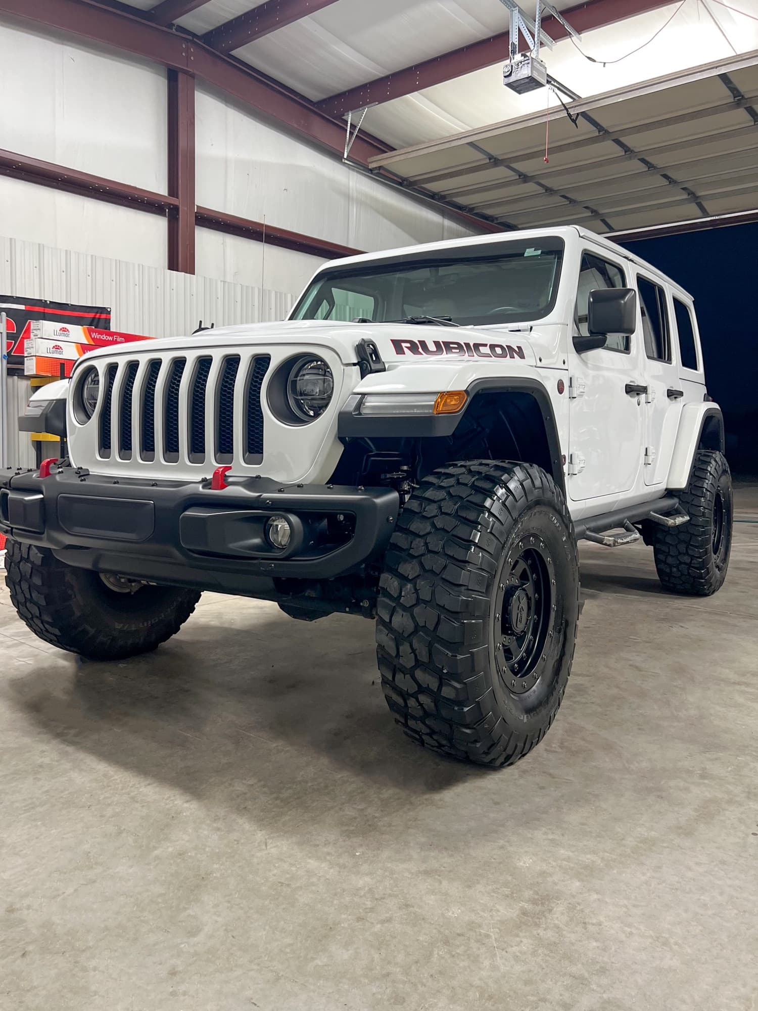 A white Jeep Wrangler Rubicon with black wheels and off-road tires inside a shop