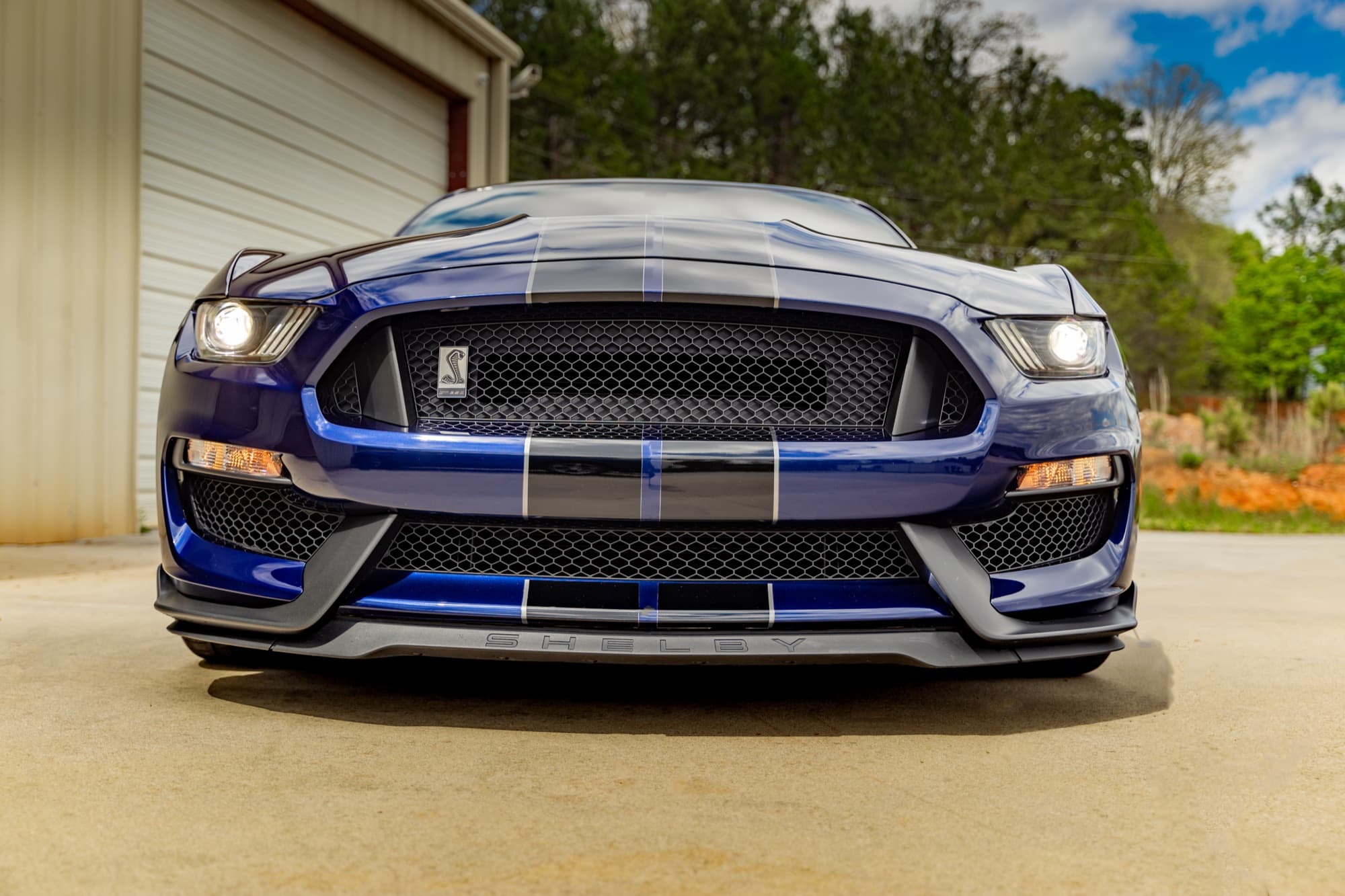 Low front view of a blue Shelby Mustang with white stripes parked outside the shop
