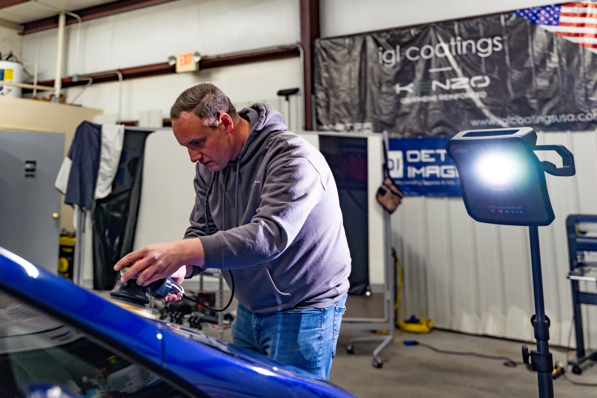A detailer working on the paint of a blue car under shop inspection lights